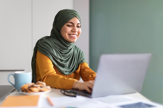 Online onderwijs Glimlachende jonge moslimvrouw in hijab die laptop thuis gebruikt
