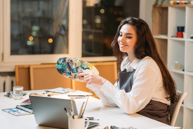 Online onderwijs Vrouwelijke artiest Kleurenreeks Kunstacademie Schilderblog Gelukkig geïnspireerde vrouw op zoek naar laptop met palet met verfspatten in licht studio-interieur