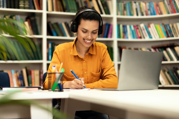 Online onderwijsconcept Glimlachende dame van gemengd ras in hoofdtelefoon die op afstand studeert in bibliotheekinterieur