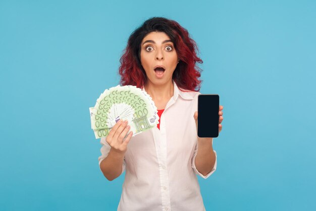 Online payment. Astonished surprised woman with fancy red hair holding cell phone and euro banknotes, shocked by money cashback and mobile application. indoor studio shot isolated on blue background