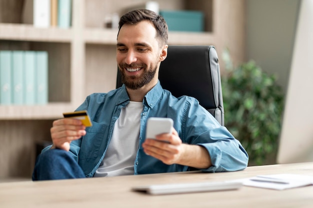 Online payment smiling young businessman using smartphone and credit card in office