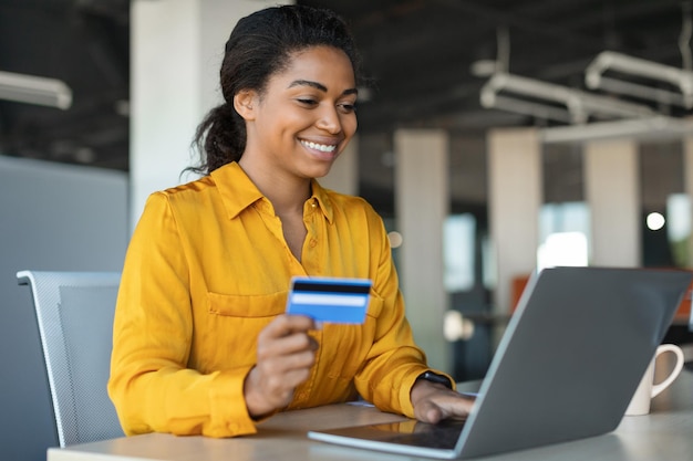 Онлайн-платежи Позитивная деловая женщина с ноутбуком и кредитной картой сидит за столом в офисном интерьере