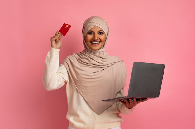 Online payments smiling muslim woman in hijab using credit card and laptop