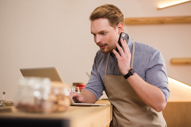 Online. Pleasant young man talking on phone while using laptop and working in the cafe