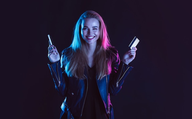 Online purchase Portrait of young beautiful woman with card phone in neon light on dark