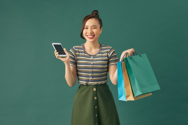 Acquisti online. bella giovane donna che tiene le borse della spesa e usa il suo smartphone con un sorriso mentre sta in piedi su sfondo verde