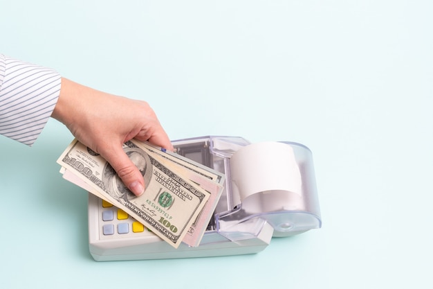 Online shopping concept. Close-up of a female hand holding several hundred dollar bills above the cash register to pay for a product or service on a blue background, top view, copy space.