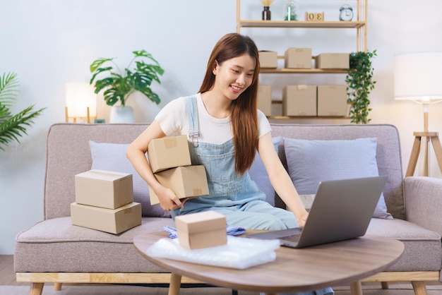 Online shopping concept Female entrepreneur holds parcel boxes and checks online orders on laptop