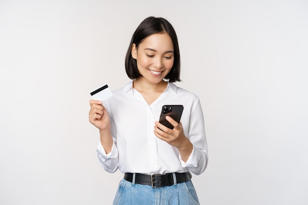 Online shopping concept Korean woman holding credit card and looking at smartphone app buying order delivery in mobile phone application standing over white background