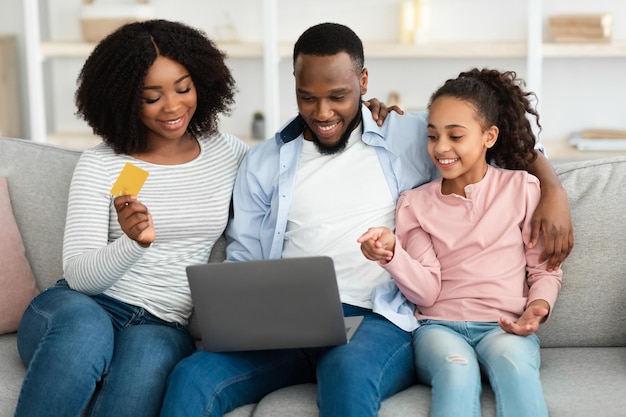 Online Shopping Concept. Portrait of cheerful African American family of three using laptop sitting on couch, teenage girl pointing at screen, showing product in online webstore, mum holding bank card