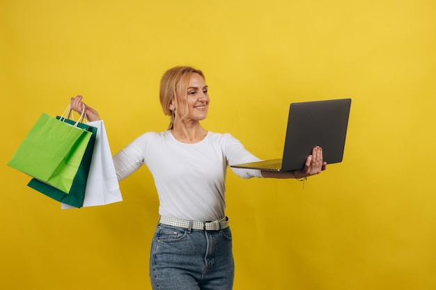 Online shopping concept portrait of happy joyful mature woman with laptop hold shopping bags isolated on yellow studio background Excited Female Enjoying Discount