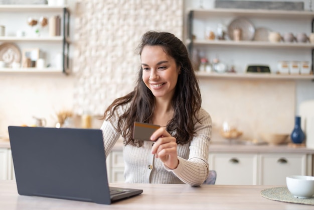 Concetto di shopping online donna positiva che utilizza carta di credito e computer portatile seduto al tavolo