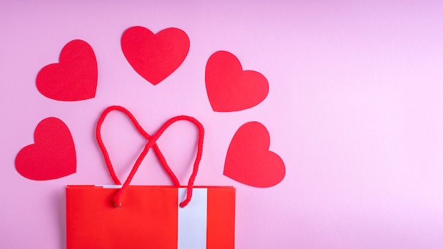 Online shopping concept. Red gift shopping bag, and red paper hearts on pink background.