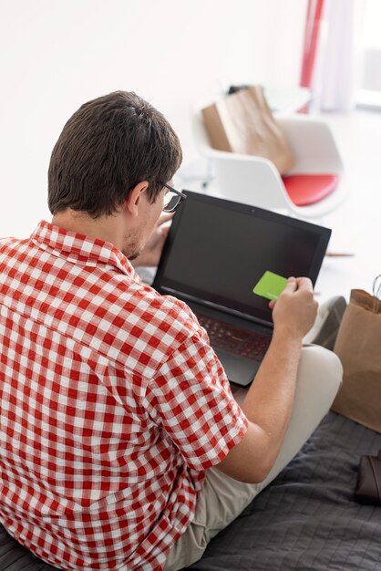 Photo online shopping concept. young man sitting on the bed and shopping online using laptop looking at the creadit card, blank screen