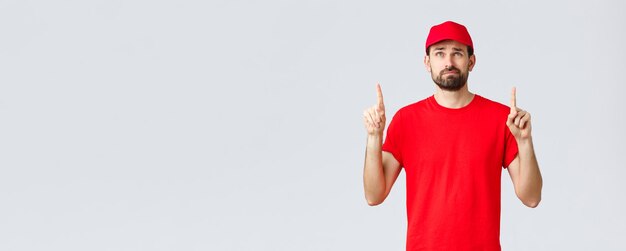 Online shopping delivery during quarantine and takeaway concept Displeased reluctant young courier employee in red uniform cap and tshirt smirk uncertain reading bad news upwards point up