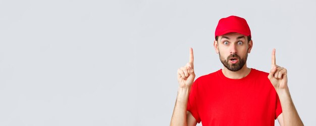 Online shopping delivery during quarantine and takeaway concept Surprised and impressed delivery guy courier in red tshirt and cap pointing fingers up gasping amazed read interesting info