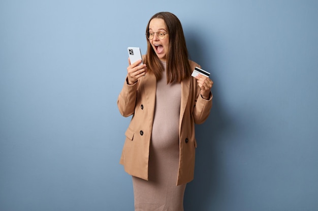Foto shopping online eccitata felice donna incinta che indossa giacca e occhiali isolata su uno sfondo blu urlando di felicità con carta di credito e cellulare in mano