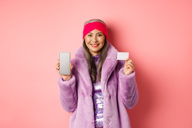 Online shopping and fashion concept. Fashionable asian senior woman showing black screen of smartphone and plastic credit card, smiling happy, pink background