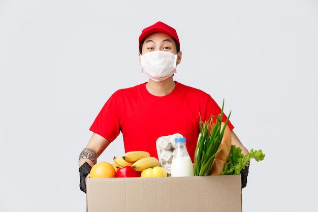 Online shopping, food delivery and coronavirus pandemic concept. Smiling delivery man in red uniform, holding box with fresh groceries, wear medical mask and gloves, contactless shopping