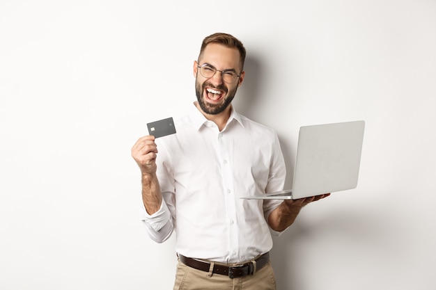 オンラインショッピング。クレジットカードを示し、白い背景の上に立って、インターネットで注文するためにラップトップを使用してハンサムな男。