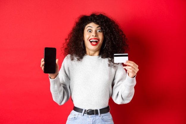 Online shopping. Happy young woman showing plastic credit card and empty phone screen, announce promo offer, standing on red wall.