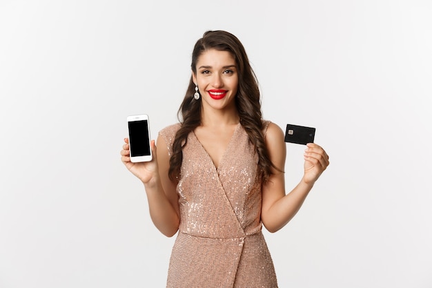 Online shopping and holidays concept. Attractive woman in party dress showing credit card and mobile screen, smiling happy, white background