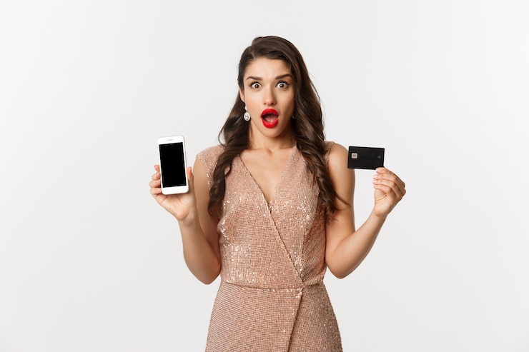 Photo online shopping and holidays concept. woman looking amazed and showing credit card with smartphone screen, standing over white background.