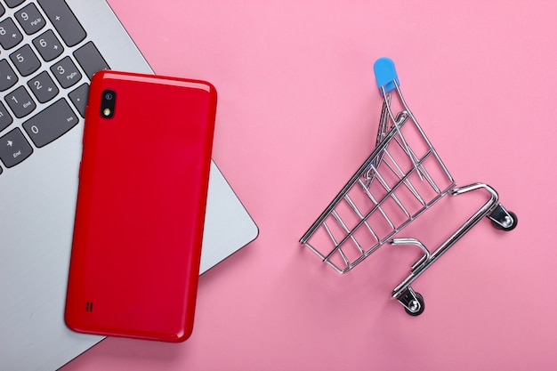 Online shopping. Laptop with smartphone and shopping trolley on a pink pastel