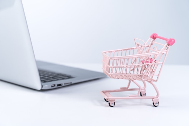 Online shopping. Mini empty pink shop cart trolley over a laptop computer on white table