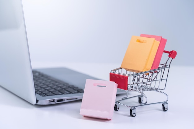 Online shopping Mini shop cart trolley with colorful paper bags over a laptop computer on white table background buying at home concept close up