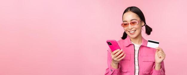 Online shopping Smiling asian girl shopper holding smartphone and credit card paying in mobile app standing over pink background