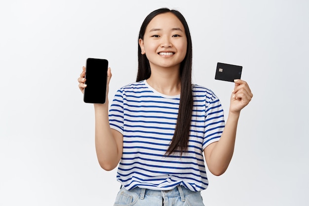 Online shopping. Smiling asian woman showing smartphone screen and credit card, recommending payment app, paying in app on white.