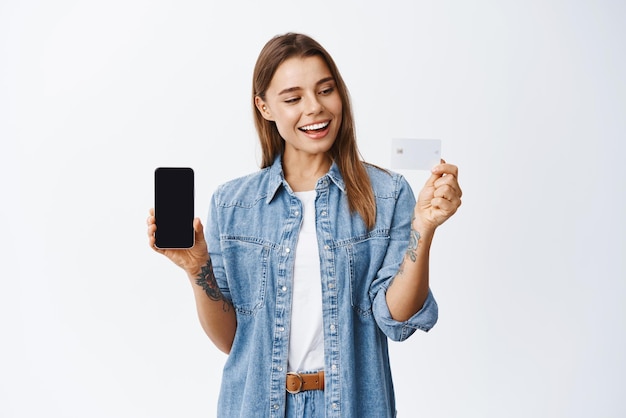 Online shopping Smiling millennial female model showing empty smartphone screen with plastic credit card mobile banking app conept white background