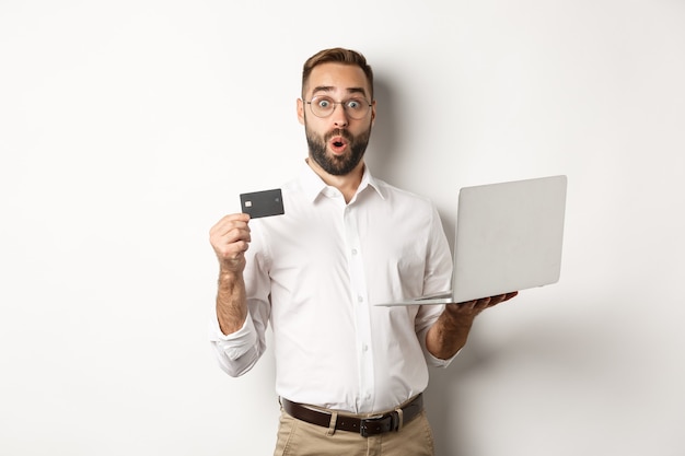 Online shopping. Surprised man holding laptop and credit card, shop internet store, standing over white background.