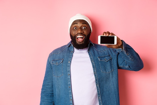 Online shopping and technology concept. Amazed Black man showing phone screen horizontally and staring at camera excited, standing over pink background