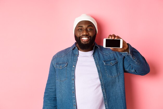 Online shopping and technology concept. Handsome Black man showing smartphone screen horizontally and smiling satisfied, recommending app, pink background