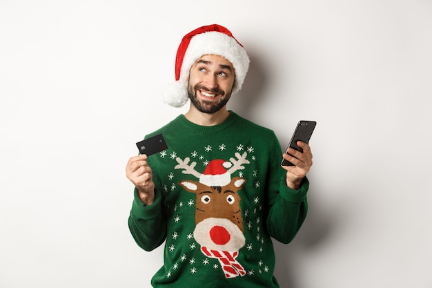 Concetto di shopping online e vacanze invernali uomo sorridente che pensa e tiene in mano una carta di credito con il cellulare...