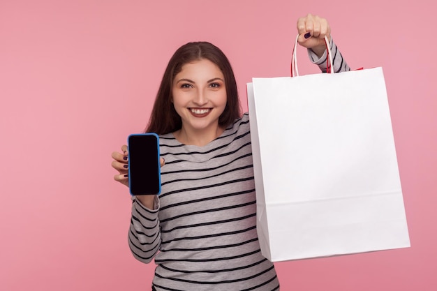 Online store app. portrait of happy woman in striped sweatshirt\
holding shopping bag and cell phone, showing mobile device with\
blank mock up for advertise. indoor studio shot, yellow\
background