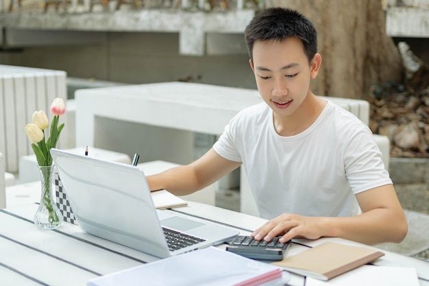 온라인 학습 개념 흰색 티셔츠를 입은 남학생은 온라인 공부를 즐기고 야외에서 새 흰색 노트북 앞에 앉아 있습니다.