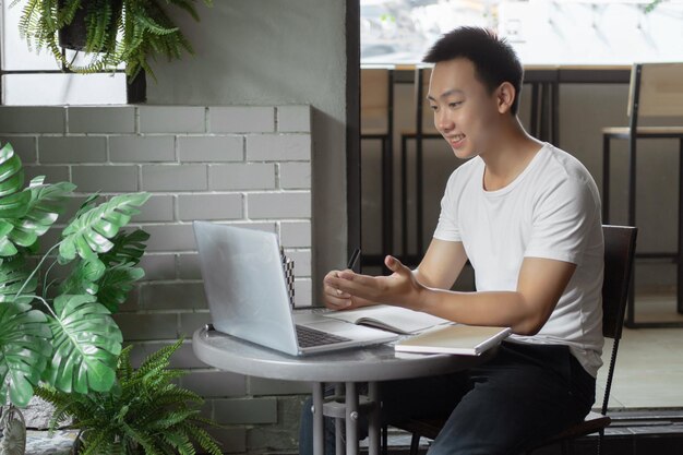 Concetto di studio online il giovane in semplice maglietta bianca e pantaloni neri che lavora usando il suo computer portatile per cercare informazioni secondo i brevi concetti scritti sul taccuino.