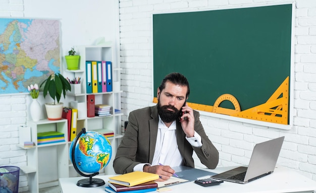 Online studying informal education male student sit in school classroom while online lesson pass the exam learning the subject serious man with beard work on laptop and use smartphone