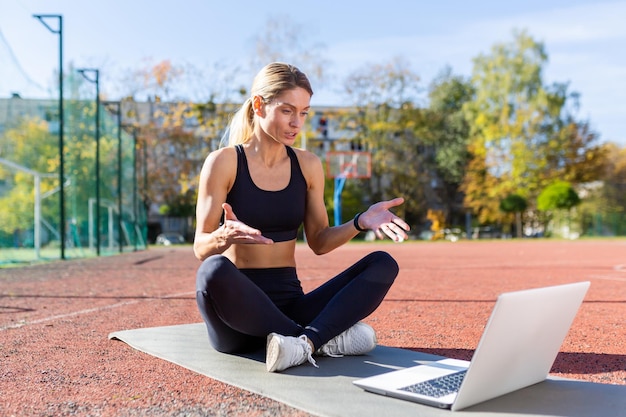 Online training female trainer sitting on sports mat in stadium at daytime using laptop for video