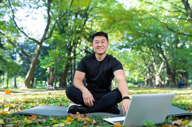 Online training handsome asian man sports trainer sits on a mat\
in the park with a laptop and