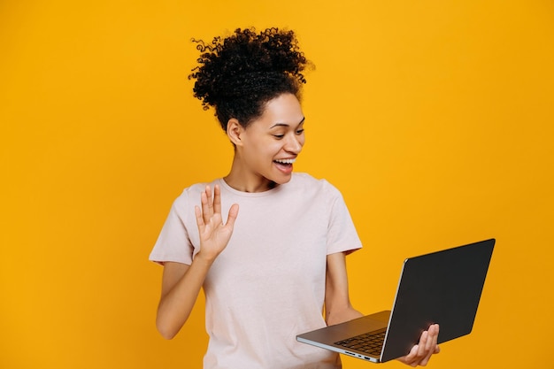 Online video communication friendly african american woman\
holding open laptop in hand doing hello gesture talking on video\
conference standing over isolated orange background smiling