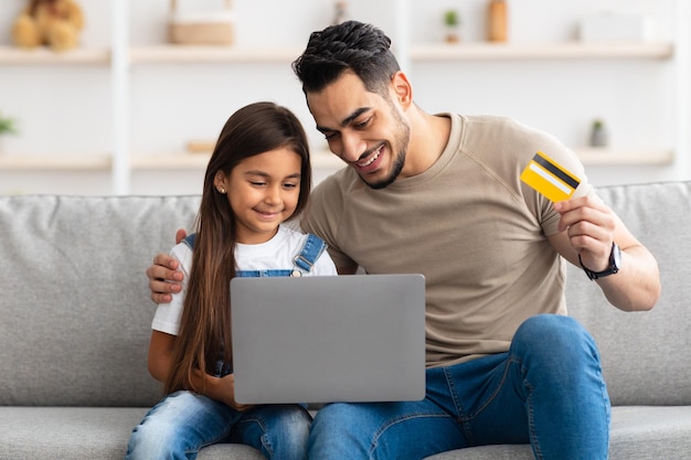 Online winkelconcept. Portret van vrolijke papa en dochter die computer gebruiken, debetcreditcard vasthouden en tonen, zittend op de bank in de woonkamer die aankopen doet op internet, kijkend naar het scherm