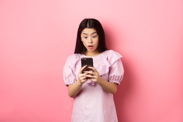 Online winkelen. aziatisch tienermeisje kijkt verbaasd naar het scherm van de mobiele telefoon, leest bericht met opgewonden uitdrukking, staande tegen roze achtergrond.