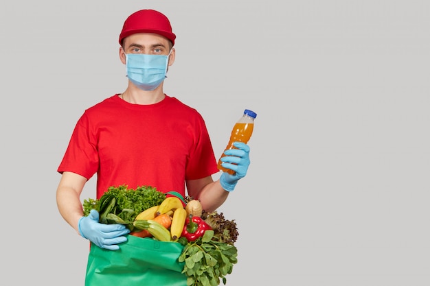 Online winkelen concept. mannelijke koerier in rood uniform, beschermend masker en handschoenen met een boodschappen doos met vers fruit en groenten. thuisbezorgd voedsel tijdens quarantaine