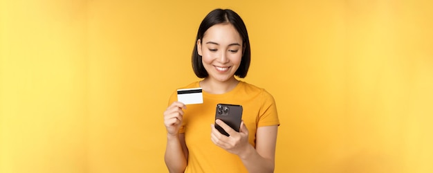 Online winkelen Glimlachend Aziatisch meisje met creditcard en mobiele telefoon-app die contactloze bestelling betaalt op smartphone-applicatie die op gele achtergrond staat