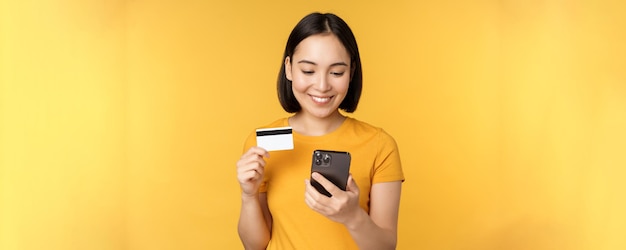 Online winkelen glimlachend aziatisch meisje met creditcard en mobiele telefoon-app die contactloze bestelling betaalt op smartphone-applicatie die op gele achtergrond staat