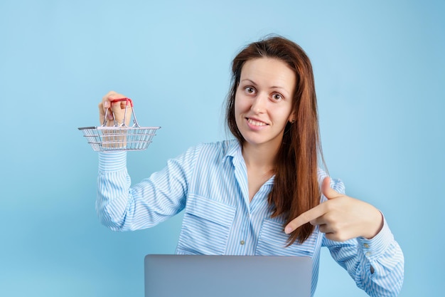 Online winkelen op internet. Een jong volwassen meisje met een laptop houdt een winkelmandje in haar handen op een blauwe achtergrond